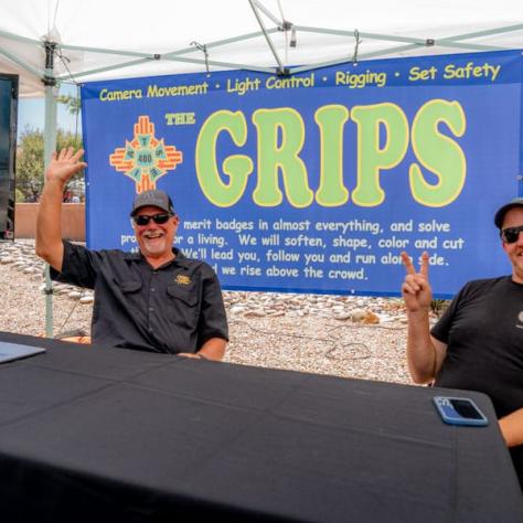 People waving hi behind table at outdoor booth. 