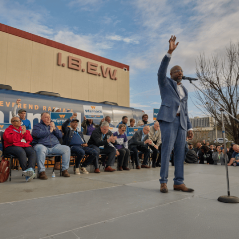 Man on stage speaking into mic with hand in the air, people sitting behind him. 