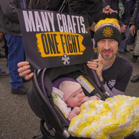 Man with baby posing for photo holding Many Crafts One Fight sign. 