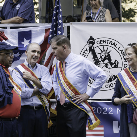 People standing wearing sashes talking to each other at a parade. 