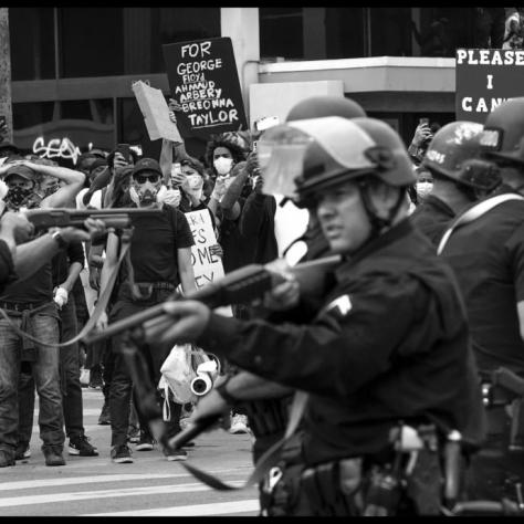 Officers pointing guns with protesters in the background. 