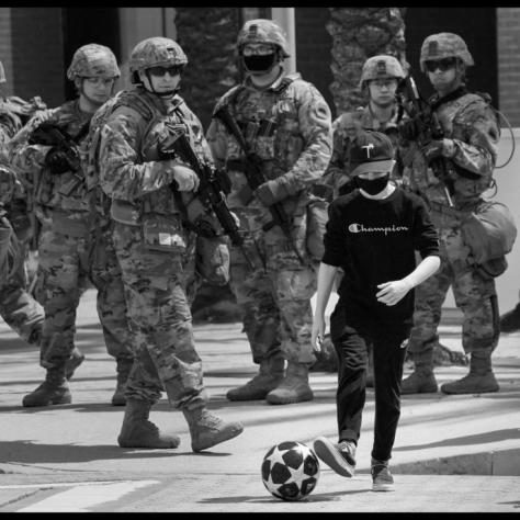 People in military outfits and guns with boy playing with soccer ball in the street. 