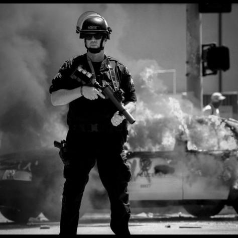 Police officer in riot gear in front of a burning car. 