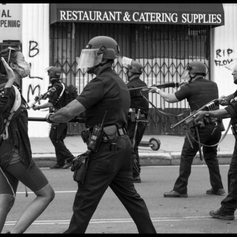 Police officers in riot gear pointing guns at protesters. 