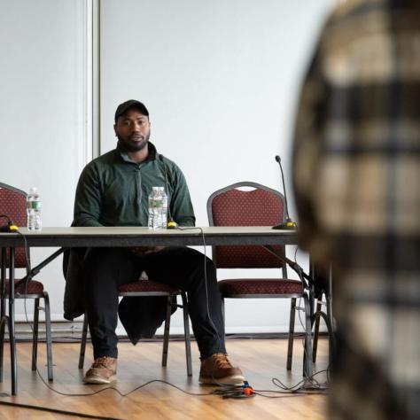 Man at front of room sitting at a table. 