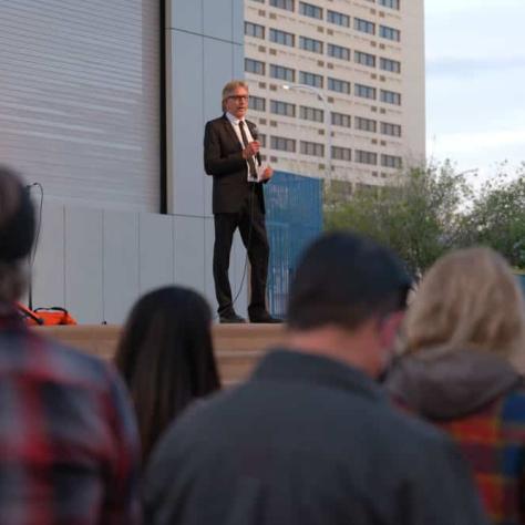 Man on stage speaking with audience watching. 