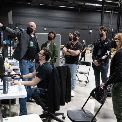 People standing around looking at a screen, man sitting in front of screen and another man is pointing at the screen. 