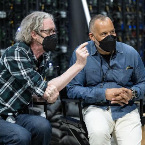 Two men wearing masks sitting in chairs talking. 