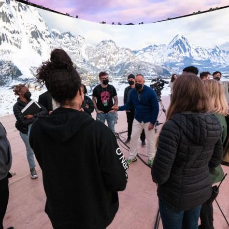 People standing listening to someone speak in front of a mountain backdrop. 