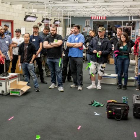 A man faces the group of people watching; colored tape is seen on the floor