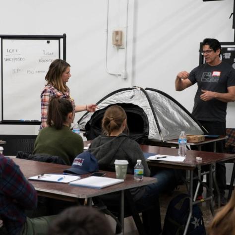 Woman shows the unzipped tent while a man speaks to the group of seated students