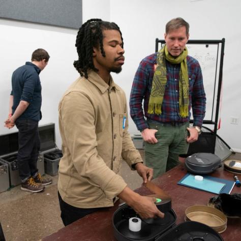 A man takes film out of a film loader while another man wearing a scarf watches